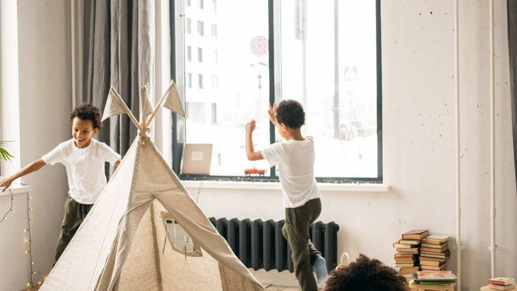 Black mother helping her daughter complete schoolwork while two younger children play nearby.